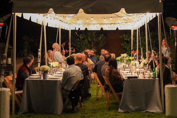 A photo of guests sitting outdoors at dinner tables underneath a canopy with twinkly lights above