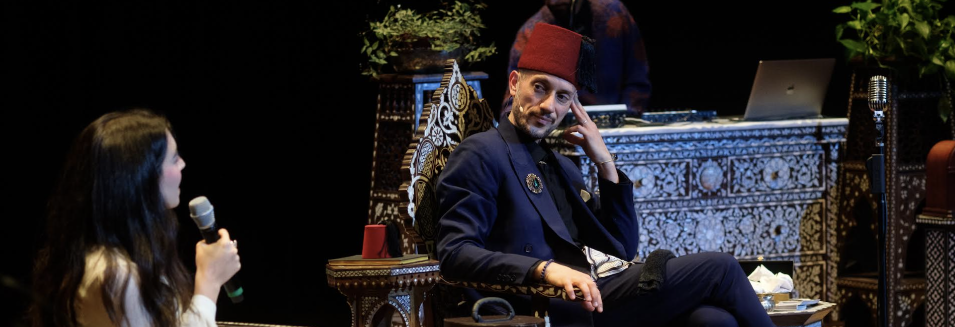 Muslim Narrative Change Fellow Omar Offendum wearing a red fez and sitting in an intricately carved wooden chair on stage with Pillars Managing Director Arij Mikati