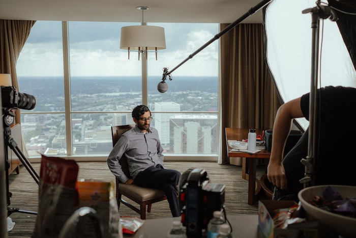 A photo of Pillars Artist Fellow Imran J. Khan being interviewed in a hotel suite, surrounded by video cameras and microphones