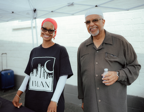 A photo of Imam Plemon El-Amin and a woman wearing a pink hijab and sunglasses smiling and standing under a white canopy outside of IMAN Atlanta's Green ReEntry apartments