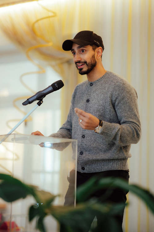A photo of Riz Ahmed wearing a black baseball cap and gray sweater speaking to a group in front of a podium