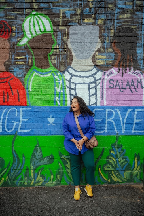 A photo of Aysha Ahmed, Development Director at Emgage, smiling in front of a colorful outdoor mural